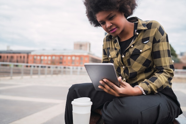 Porträt der jungen afroamerikanischen lateinamerikanischen Frau, die ihr digitales Tablett beim Sitzen auf einer Bank im Freien verwendet. Technologiekonzept.