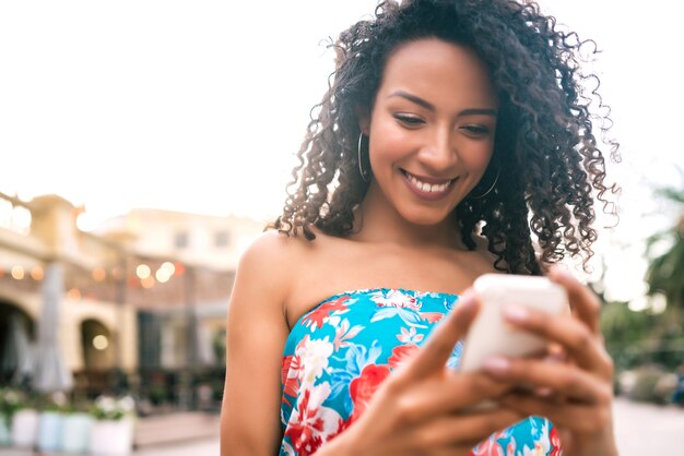 Porträt der jungen afroamerikanischen lateinamerikanischen Frau, die ihr Handy draußen in der Straße benutzt. Technologiekonzept.