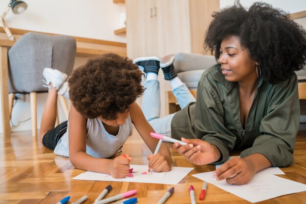 Porträt der jungen afroamerikanischen Mutter und des Sohnes, die mit Buntstiften auf warmem Boden zu Hause zeichnen. Familienkonzept.