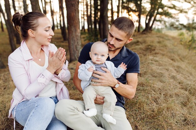 Porträt der jungen attraktiven Familie mit dem kleinen kleinen Sohn, der im schönen Herbstkiefernwald am sonnigen Tag aufwirft. Hübscher Mann und seine hübsche brünette Frau