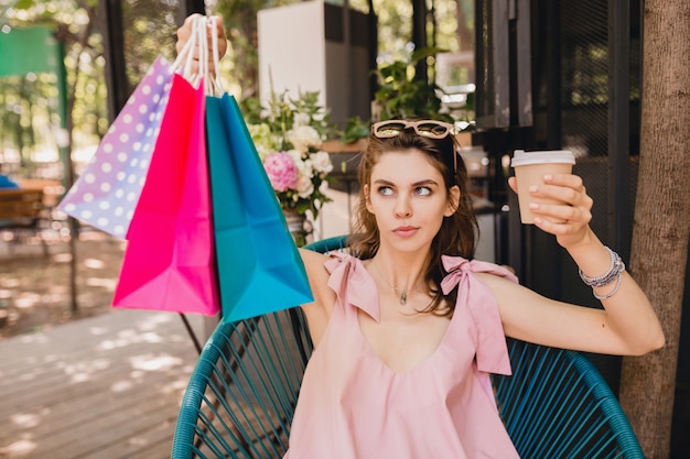 Porträt der jungen attraktiven Frau, die im Café mit Einkaufstüten sitzt, die Kaffee trinken, Sommermode-Outfit, rosa Baumwollkleid, trendige Kleidung, verwirrt, denkend