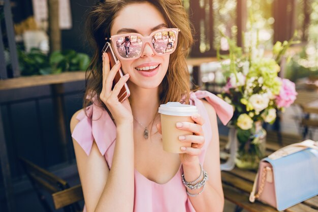 Porträt der jungen attraktiven Frau, die im Café sitzt, Sommermode-Outfit, rosa Baumwollkleid, Sonnenbrille, lächelnd, Kaffee trinkend, stilvolle Accessoires, trendige Kleidung, am Telefon sprechend