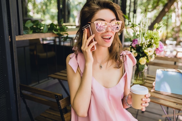 Porträt der jungen attraktiven Frau, die im Café sitzt, Sommermode-Outfit, rosa Baumwollkleid, Sonnenbrille, lächelnd, Kaffee trinkend, stilvolle Accessoires, trendige Kleidung, am Telefon sprechend