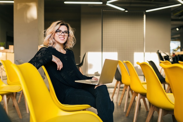 Porträt der jungen attraktiven Frau, die im Hörsaal sitzt, der auf Laptop trägt, der Brille trägt, Student, der im Klassenzimmer mit vielen gelben Stühlen lernt
