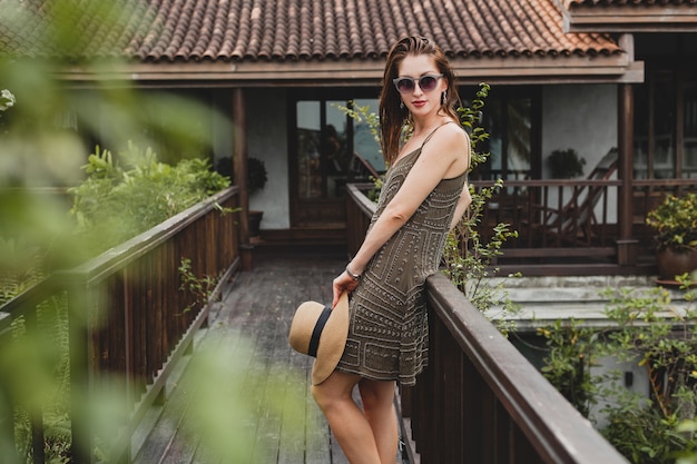 Porträt der jungen attraktiven Frau im eleganten Kleid, im Strohhut, im Sommerstil, im Modetrend, im Urlaub, im Lächeln, im stilvollen Zubehör, in der Sonnenbrille, im Aufstellen auf tropischer Villa auf Bali