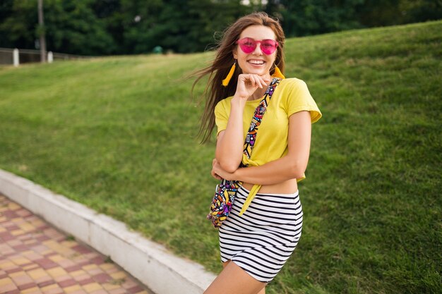 Porträt der jungen attraktiven stilvollen Frau, die im Stadtpark, lächelnde fröhliche Stimmung, positiv, tragendes gelbes Oberteil, gestreiften Minirock, Handtasche, rosa Sonnenbrille, Sommerart-Modetrend aufwirft