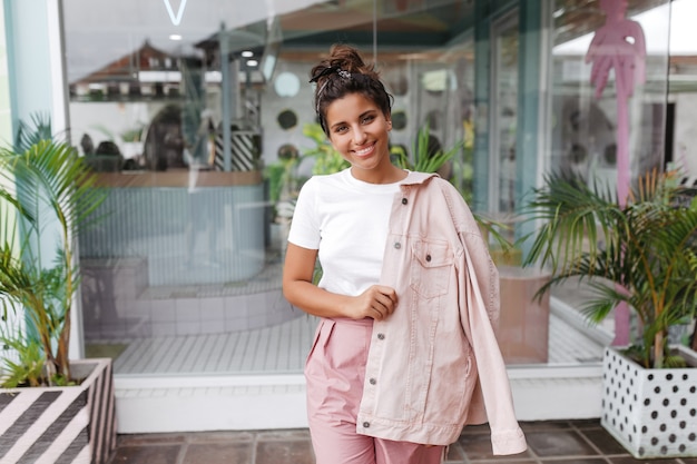 Porträt der jungen brünetten Frau im weißen T-Shirt und in der rosa Jeansjacke, die mit Lächeln neben niedlicher Bar aufwirft