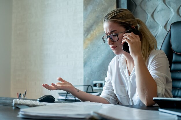 Porträt der jungen Büroangestelltenfrau, die am Schreibtisch mit Dokumenten sitzt, die auf Handy nervös und gestresst arbeiten im Büro sprechen