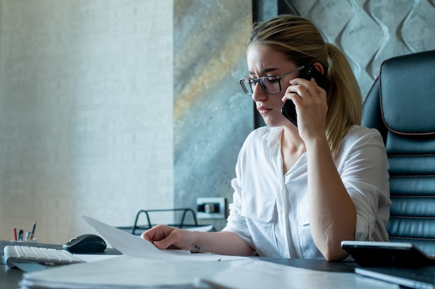 Porträt der jungen Büroangestelltenfrau, die am Schreibtisch mit Dokumenten sitzt, die auf Handy nervös und gestresst arbeiten im Büro sprechen