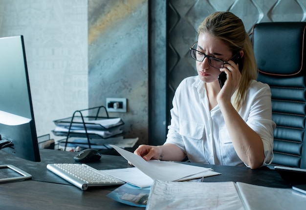 Kostenloses Foto porträt der jungen büroangestelltenfrau, die am schreibtisch mit dokumenten sitzt, die auf handy nervös und gestresst arbeiten im büro sprechen