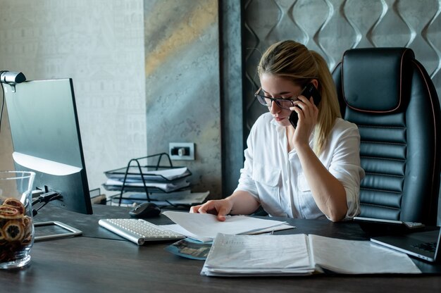 Porträt der jungen Büroangestelltenfrau, die am Schreibtisch mit Dokumenten sitzt, die auf Mobiltelefon mit sicherem und ernstem Ausdruck auf Gesicht sprechen, das im Büro arbeitet