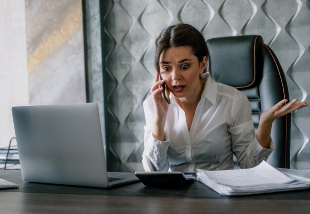 Porträt der jungen Büroangestelltenfrau, die am Schreibtisch mit Dokumenten sitzt, die auf Mobiltelefon mit wütendem Ausdruck frustrierten nervösen und gestressten Arbeiten im Büro sprechen