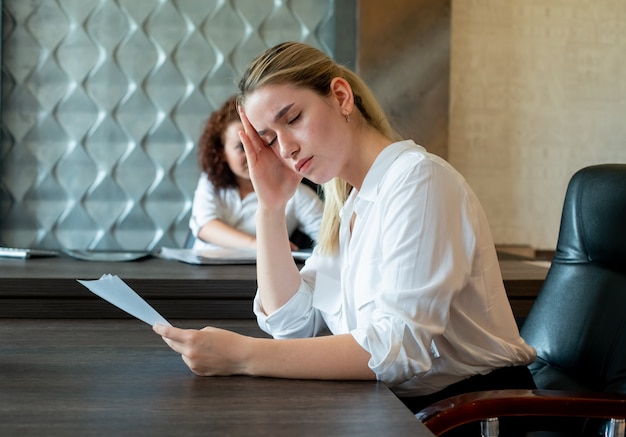 Porträt der jungen Büroangestelltenfrau, die am Schreibtisch mit Dokumenten sitzt, die gelangweilt und überarbeitet arbeiten, die im Büro sitzen