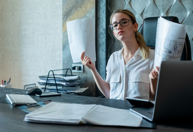 Porträt der jungen Büroangestelltenfrau, die am Schreibtisch mit Dokumenten sitzt, die Kamera mit ernstem sicherem Ausdruck auf Gesicht betrachten, das im Büro arbeitet