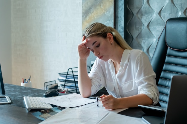 Porträt der jungen Büroangestelltenfrau, die am Schreibtisch mit Dokumenten sitzt, die müde und gelangweilt im Büro arbeiten