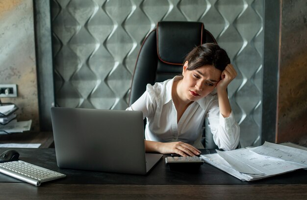 Porträt der jungen Büroangestelltenfrau, die am Schreibtisch mit Dokumenten und Laptop-Computer sitzt, die müde und überarbeitetes Arbeitsprozessbürokonzept suchen