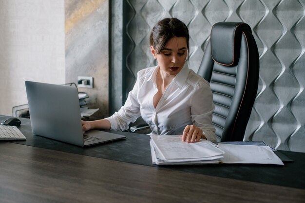 Porträt der jungen Büroangestelltenfrau, die am Schreibtisch mit Dokumenten unter Verwendung des Laptop-Computers sitzt, der zuversichtlich und beschäftigt im Büro arbeitet
