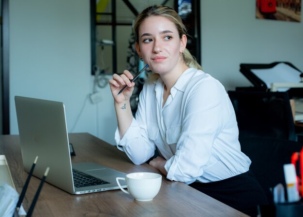 Porträt der jungen büroangestelltenfrau, die am schreibtisch mit laptop-computer sitzt und beiseite denkt mit nachdenklichem ausdruck, der im büro arbeitet