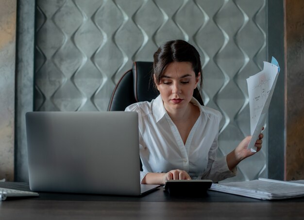 Porträt der jungen Büroangestelltenfrau, die am Schreibtisch mit Laptop und Dokumenten rechnet, die mit ernsthaftem Ausdruck beschäftigt und zuversichtlich arbeiten Prozessbürokonzept berechnen