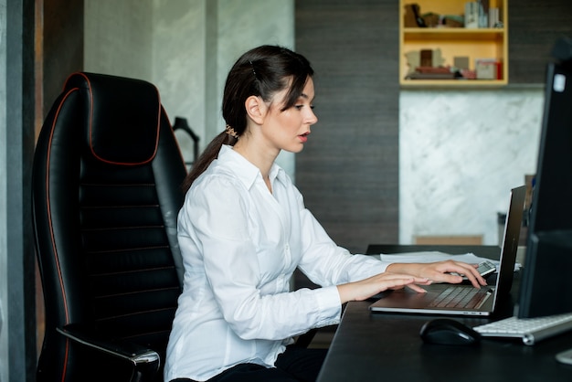 Porträt der jungen Büroangestelltenfrau, die am Schreibtisch sitzt unter Verwendung des Laptop-Computers, der beschäftigt mit zuversichtlichem ernstem Ausdruck auf Gesicht arbeitet, das im Büro arbeitet