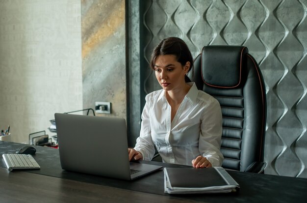 Porträt der jungen Büroangestelltenfrau, die am Schreibtisch unter Verwendung des Laptop-Computers sitzt, der beschäftigt mit ernstem und sicherem Ausdruck auf Gesicht arbeitet, das im Büro arbeitet