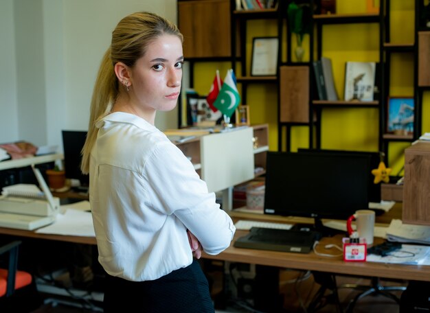Porträt der jungen Büroangestelltenfrau, die seitlich mit verschränkten Armen steht und Kamera mit ernstem Gesicht unzufrieden und sehr besorgt im Amt betrachtet
