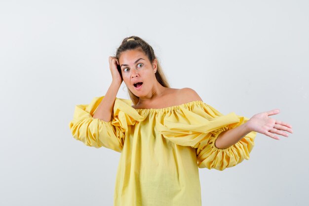 Porträt der jungen Dame, die etwas zeigt, Hand auf Kopf im gelben Kleid hält und erstaunte Vorderansicht schaut