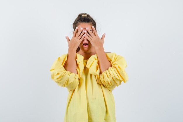 Porträt der jungen Dame, die Hände auf Augen im gelben Kleid hält und aufgeregte Vorderansicht schaut