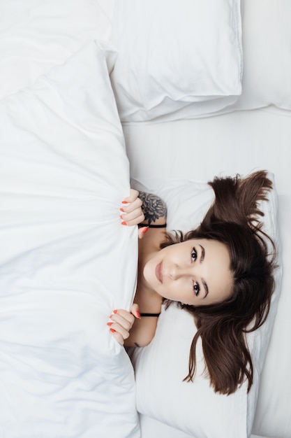 Porträt der jungen Dame, die im Bett unter weißer Bettdecke liegt und ihren Körper bedeckt