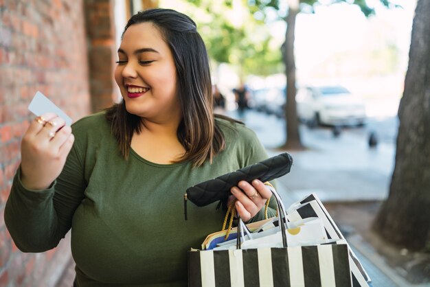 Porträt der jungen Frau der Übergröße, die eine Kreditkarte und Einkaufstaschen draußen auf der Straße hält. Einkaufs- und Verkaufskonzept.