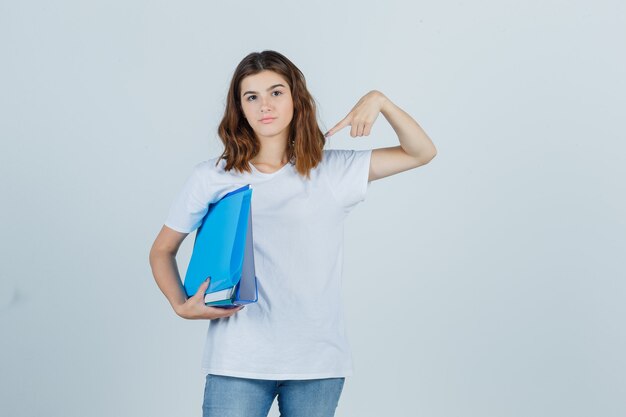 Porträt der jungen Frau, die auf Ordner im weißen T-Shirt, in den Jeans und in der selbstbewussten Vorderansicht zeigt