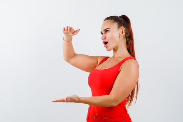 Porträt der jungen Frau, die Größenzeichen im roten Trägershirt, in der Hose und in der verwirrten Vorderansicht zeigt