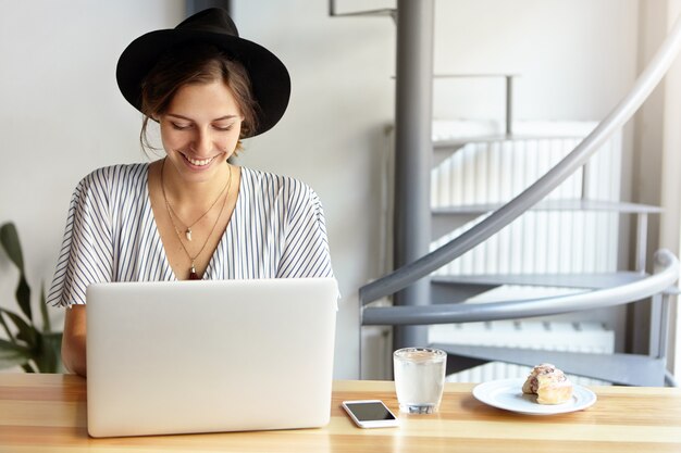 Porträt der jungen Frau, die großen Hut trägt und Laptop verwendet