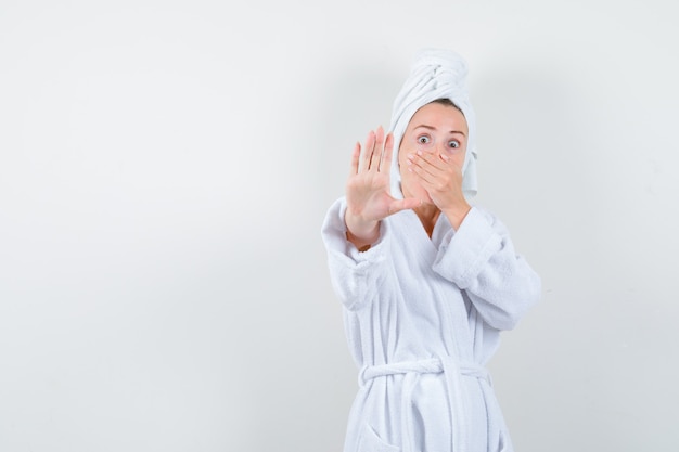 Porträt der jungen Frau, die Hand auf Mund hält, Stoppgeste in weißem Bademantel, Handtuch zeigt und ängstliche Vorderansicht schaut