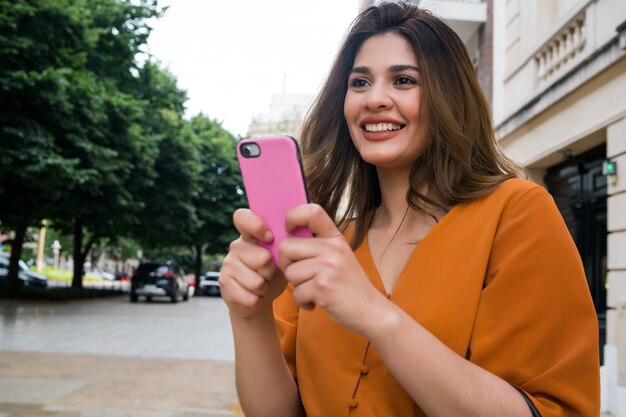 Porträt der jungen Frau, die ihr Handy benutzt, während sie draußen auf der Straße geht