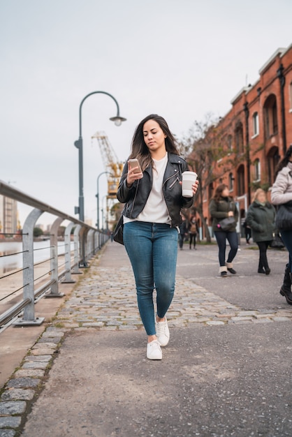 Porträt der jungen Frau, die ihr Handy benutzt, während sie mit Tasse Kaffee geht. Stadt- und Kommunikationskonzept.