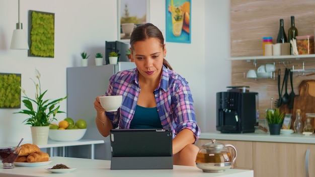 Porträt der jungen Frau, die morgens Tablette verwendet, die am Tisch in der Küche sitzt und Tee trinkt. Arbeiten von zu Hause aus mit einem Gerät mit Internettechnologie, Tippen, auf Gadget während des Frühstücks.