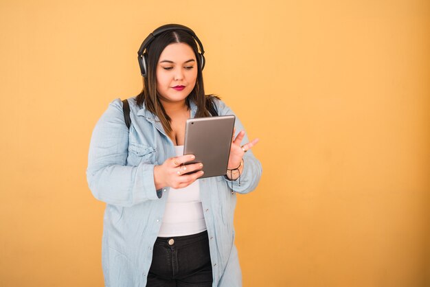 Porträt der jungen Frau, die Musik mit Kopfhörern und digitalem Tablett draußen gegen gelbe Wand hört