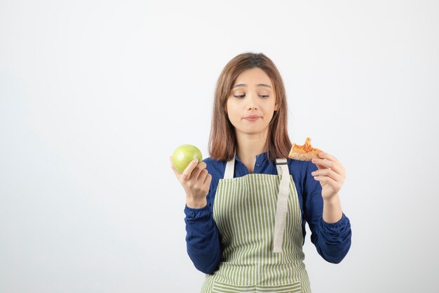 Porträt der jungen Frau, die Scheibe Pizza und Apfel über weißer Wand betrachtet.