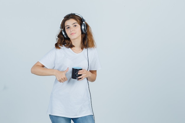 Porträt der jungen Frau, die Tasse des Getränks hält, zeigt Daumen oben im weißen T-Shirt und schaut erfreute Vorderansicht