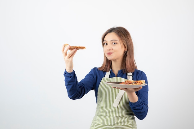 Porträt der jungen Frau in Schürze, die mit Pizza auf Weiß posiert