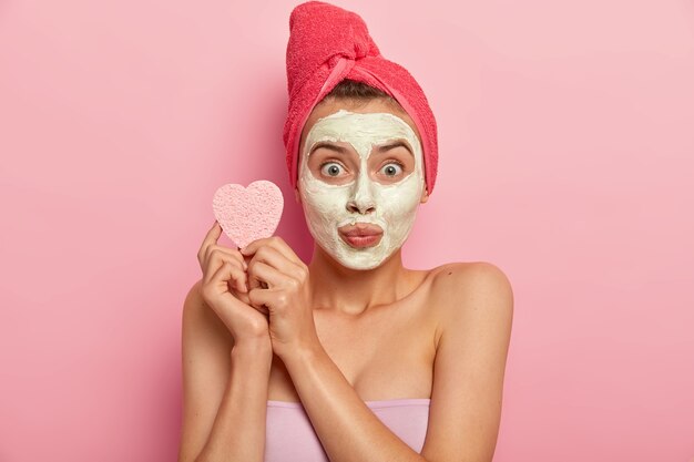 Porträt der jungen Frau mit mineralischer natürlicher Maske im Gesicht, hat tägliche Hautpflegebehandlung, hält kosmetischen Schwamm zum Abwischen des Teints, reinigt die Haut und entfernt abgestorbene Hautzellen, hat Bad zu Hause