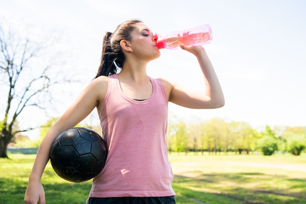 Porträt der jungen Fußballspielerin, die Pause auf dem Spielfeld und Trinkwasser aus der Flasche macht