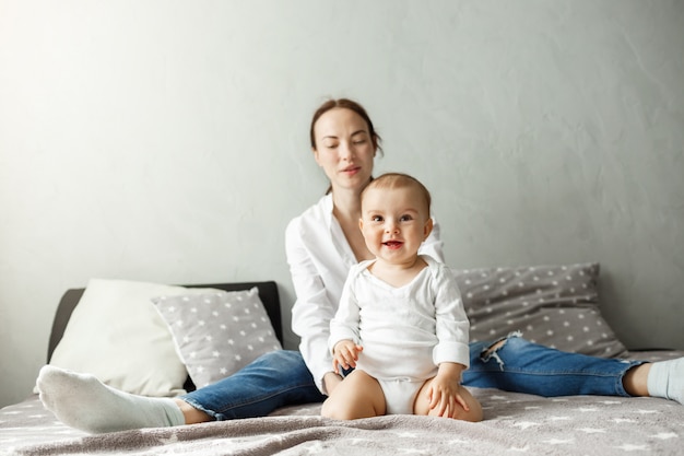 Porträt der jungen glücklichen Familie der hübschen Mutter und des neugeborenen Sohnes, die kostbare Zeit zusammen verbringen, lächelnd und im Schlafzimmer spielend.