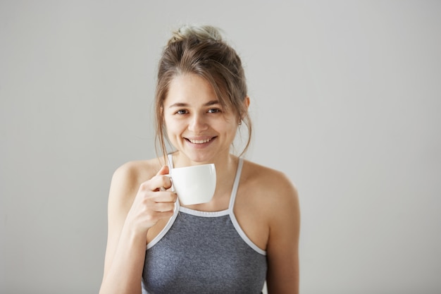 Porträt der jungen glücklichen schönen Frau lächelnd, die trinkende Tasse Kaffee am Morgen über weißer Wand hält.