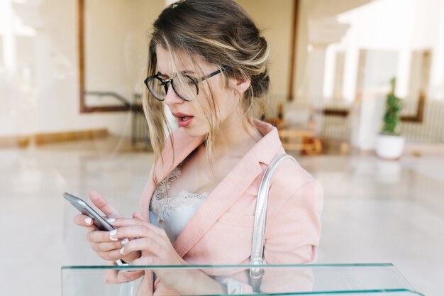 Porträt der jungen hübschen Frau, stilvolle Geschäftsdame überrascht Blick auf Smartphone, hält silbernes Telefon in Händen, in der Halle stehen. Sie hat eine weiße kurze Maniküre und trägt eine elegante rosa Jacke.