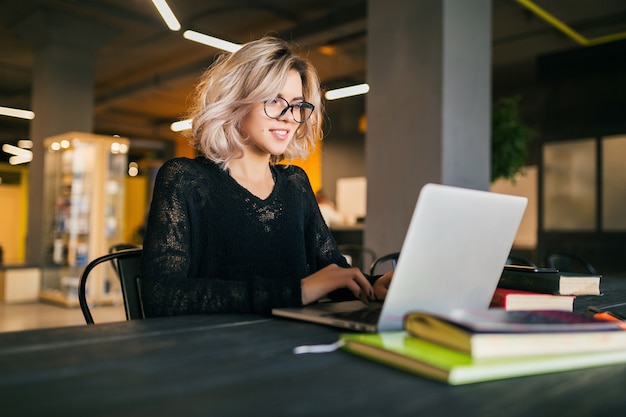 Porträt der jungen hübschen lächelnden Frau, die am Tisch im schwarzen Hemd sitzt, das am Laptop im mitarbeitenden Büro arbeitet und Brille trägt