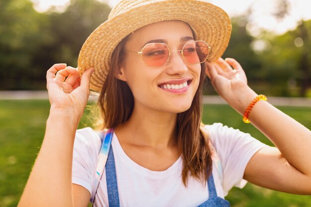 Porträt der jungen hübschen lächelnden Frau im Strohhut und in der rosa Sonnenbrille, die im Park, Sommermodeart, buntes Hipster-Outfit gehen