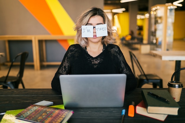 Porträt der jungen hübschen müden Frau mit Papieraufklebern auf Gläsern, die am Tisch im schwarzen Hemd arbeiten, das am Laptop im mitarbeitenden Büro arbeitet, lustiger Gesichtsausdruck, frustrierte Emotion