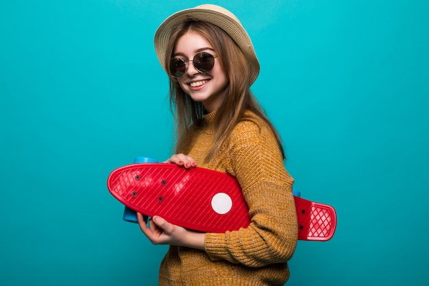 Porträt der jungen jugendlich Frau in der Sonnenbrille und im Hut, die Skateboard halten, während lokalisiert über grüner Wand stehen